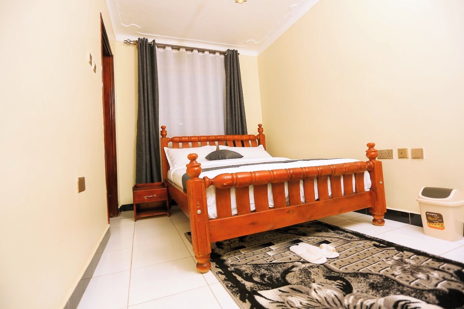 A small, neatly arranged bedroom with a wooden bed, a nightstand, a bedside table, a patterned rug, and a trash bin.