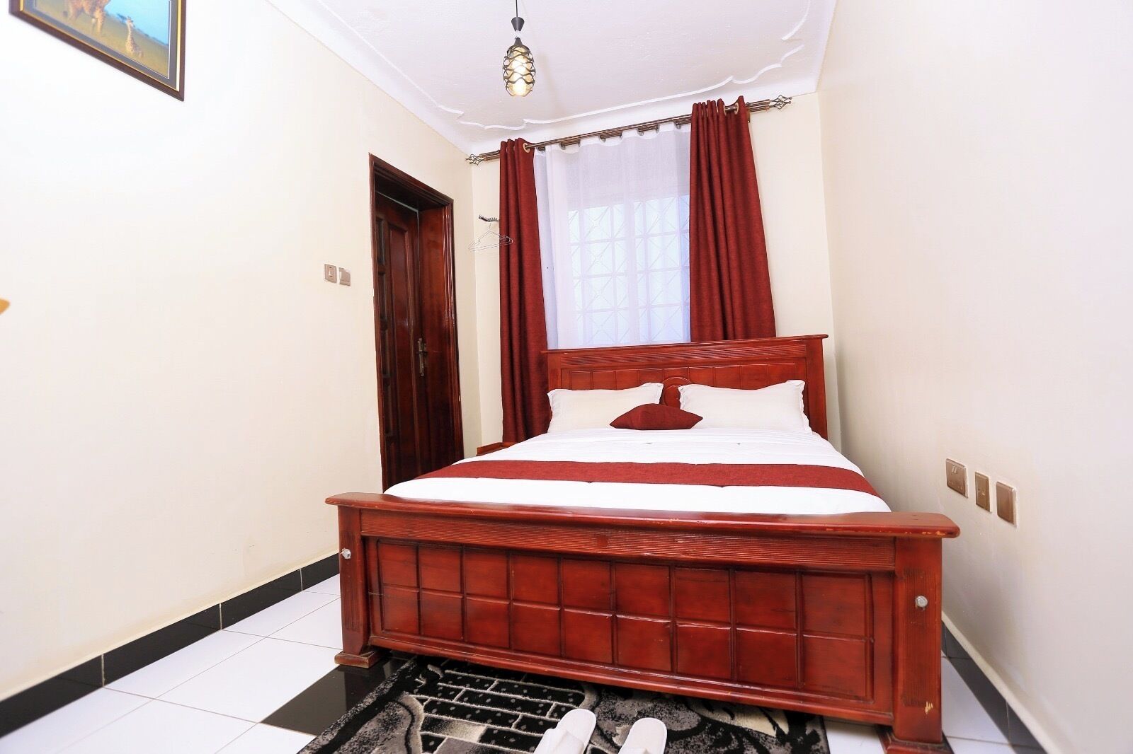 A bedroom with a wooden bed, white bedding, a window with red curtains, and a ceiling light fixture.