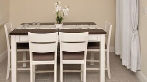 Un coin repas avec une table dressée pour deux personnes, des chaises blanches, un vase de fleurs blanches et un tableau accroché au mur.
