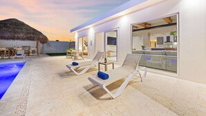 A modern outdoor lounge area with sun loungers and a pool, connected to a building with large windows and a thatched-roof structure in the background. - NEW 6BR Playground Private Pool Pro-BBQ Palapa (Noord)