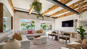 A modern living room with a grey sofa, round coffee table, wicker furniture, and a view of a pool and palm trees through large windows. - NEW 6BR Playground Private Pool Pro-BBQ Palapa (Noord)