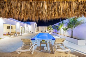 Un área junto a la piscina con sillones de descanso, una mesa y un techo de paja.