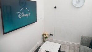 Ein modernes Wohnzimmer mit einem an der Wand montierten Flachbildfernseher, der das Disney+-Logo zeigt, einem grauen Sofa, einem weißen Couchtisch und einer Vase mit weißen Blumen.