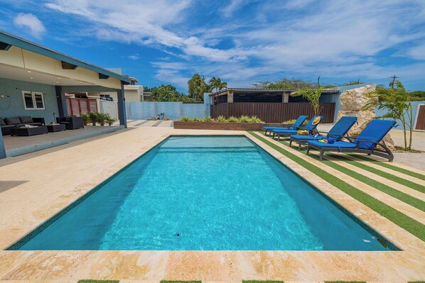 A rectangular swimming pool with blue water, surrounded by a tiled deck and a grassy area with blue lounge chairs.