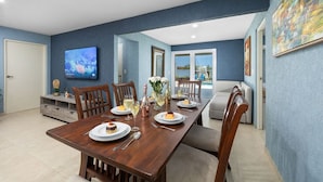 Un coin repas avec une table en bois dressée pour quatre personnes, entourée de chaises. Un téléviseur est fixé au mur, et un canapé est visible au fond.
