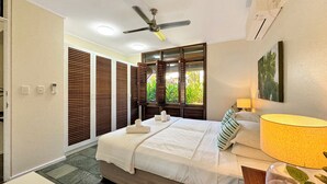 Een slaapkamer met een bed, een plafondventilator, een raam met uitzicht op groen en een wandairconditioner.