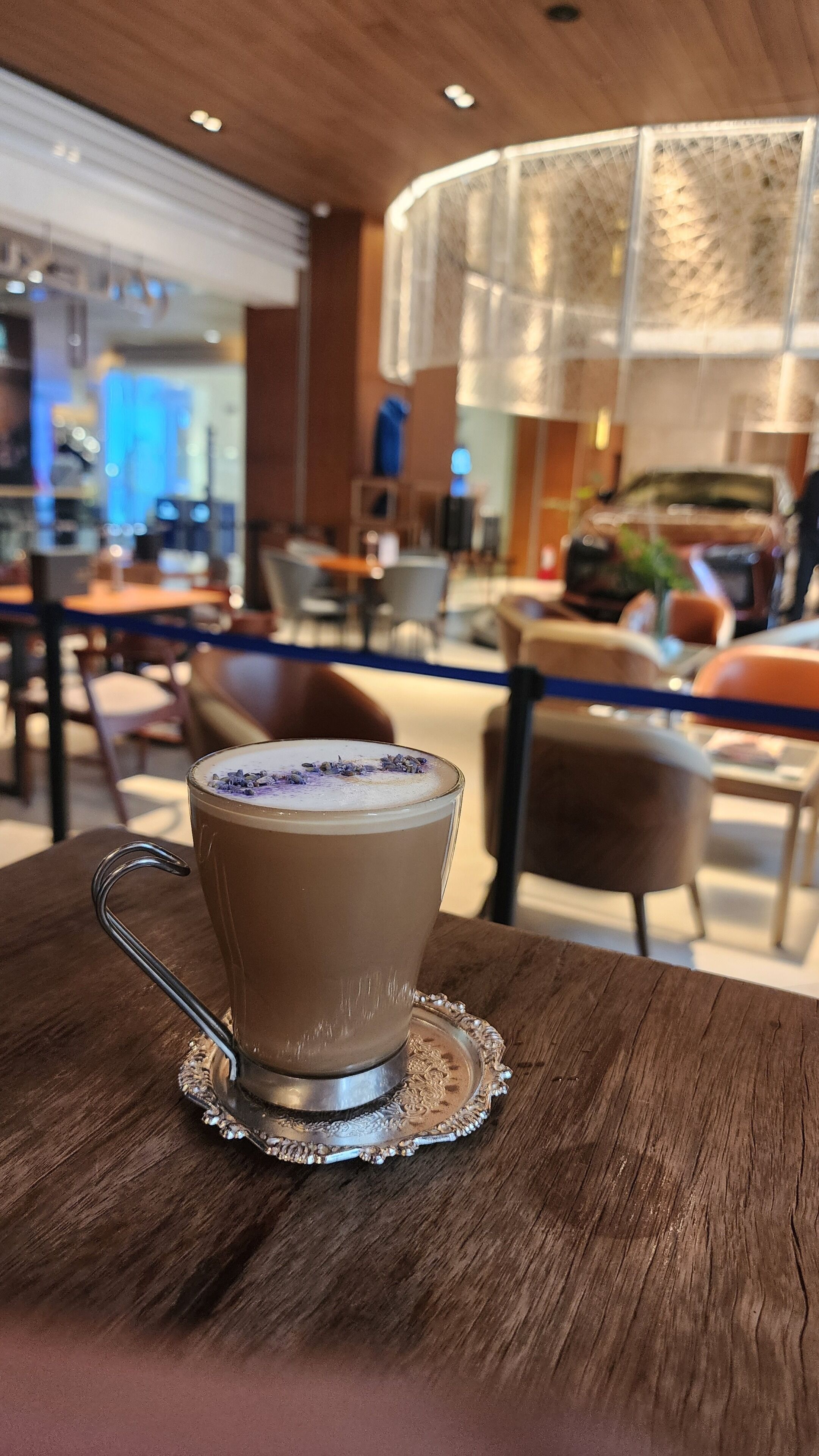 A cup of coffee on a wooden table in a cafe setting with a blurred background of chairs and tables.
