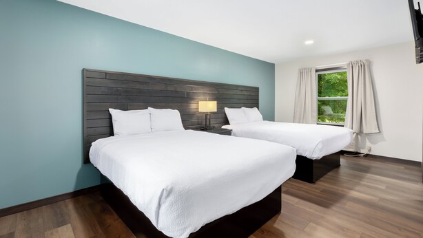 A modern hotel room with two beds, a wooden headboard, a nightstand with a lamp, a window with curtains, and a view of greenery outside. - Rustic Riverside Lodge | WiFi | Pet-Friendly (Aitkin)