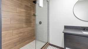 A modern bathroom with a wooden wall, a glass shower enclosure, and a dark-toned vanity with a round mirror. - Rustic Riverside Lodge | WiFi | Pet-Friendly (Aitkin)