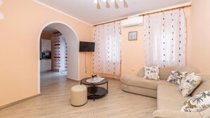 Un soggiorno con divano, poggiapiedi e TV a parete. Si intravede una cucina attraverso un arco.