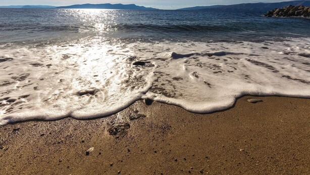 Una spiaggia con onde che lambiscono dolcemente la riva, cielo sereno e montagne in lontananza.