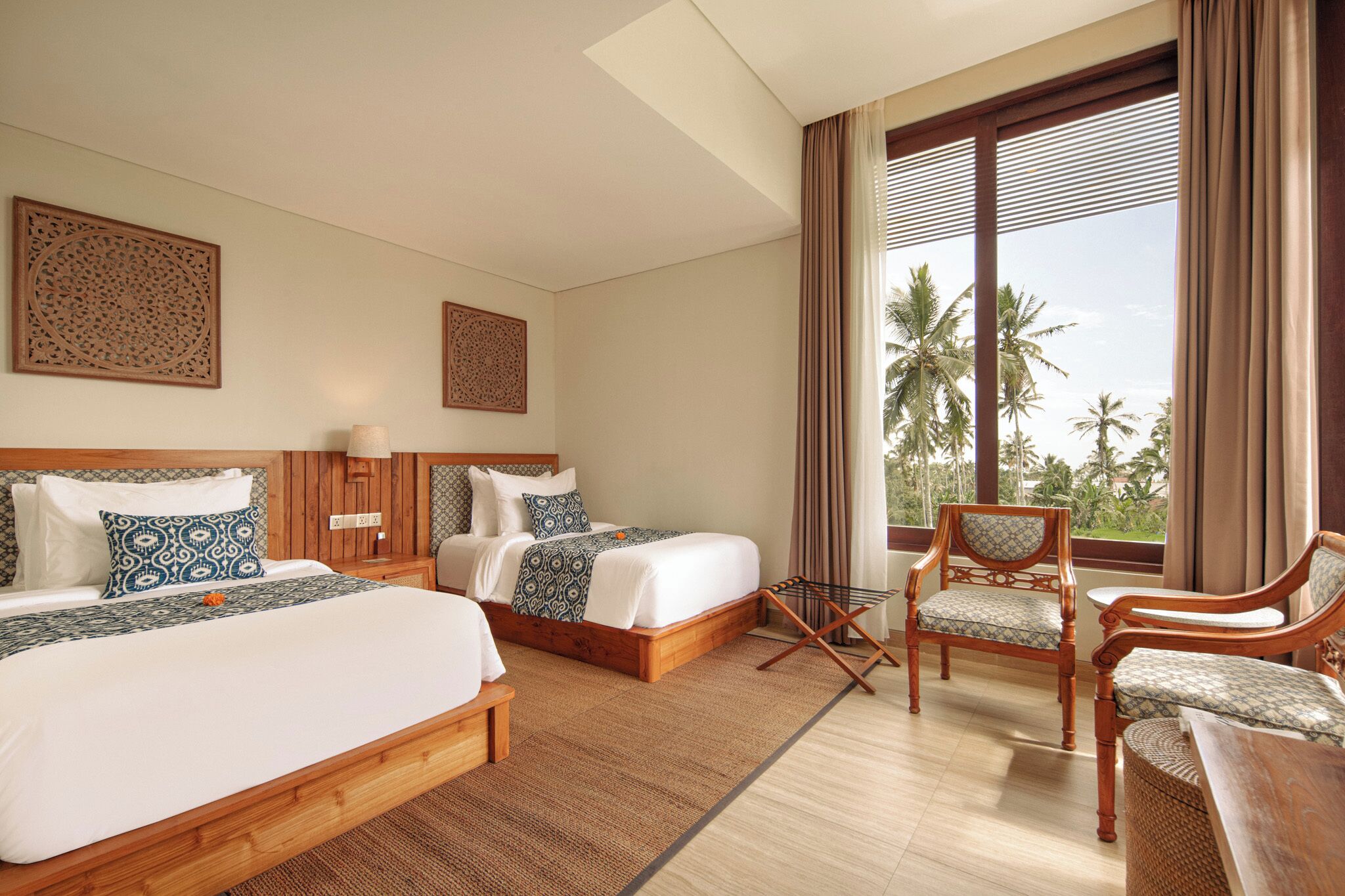 Habitación de hotel con dos camas, un área de descanso con sillas, un ventanal con cortinas y paneles decorativos en la pared.