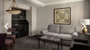 A living room with a grey sofa, a glass coffee table, and a dining area with a round table and chairs. There is a kitchen area with dark cabinets and a hanging light fixture. - The Manhattan Club (New York)