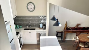 Une cuisine moderne avec des meubles blancs, un dosseret en carreaux noirs et un lave-vaisselle encastré. On y trouve un coin repas avec une table et des chaises en bois, ainsi qu’un escalier menant à un autre niveau.
