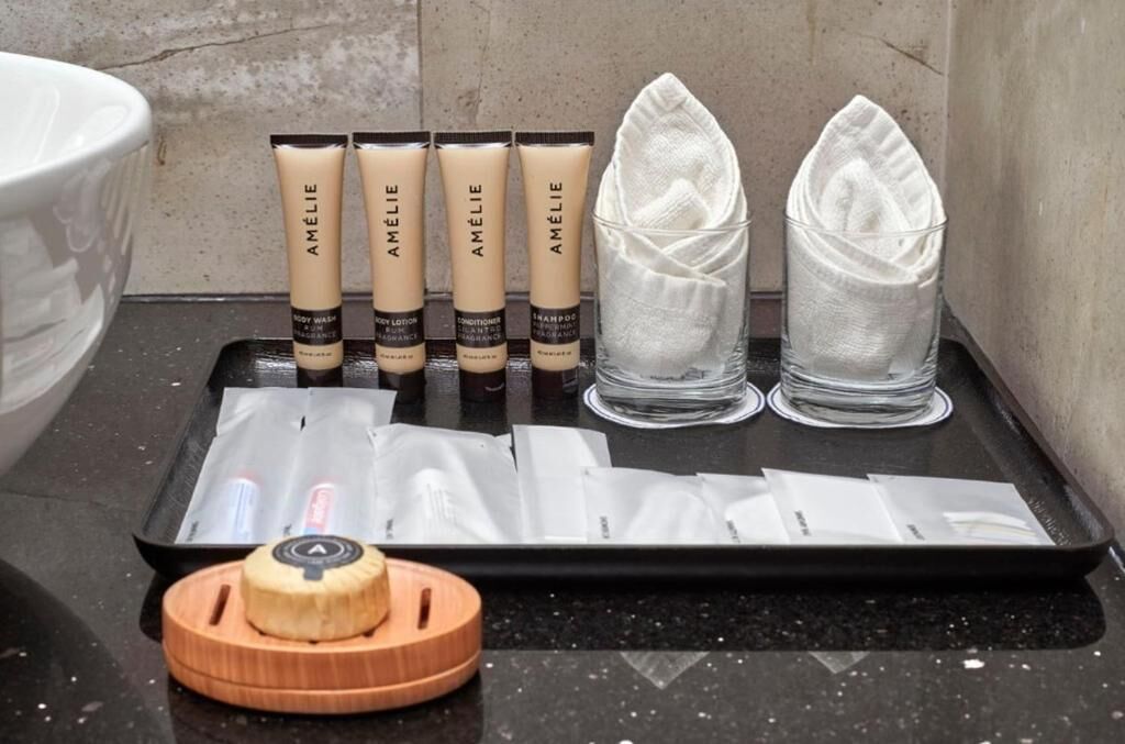 Una bandeja con productos de cuidado corporal Amelie, incluyendo loción, acondicionador y champú, junto con un recipiente de madera y una toalla doblada.