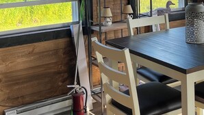 A dining area with a table and chairs, a fire extinguisher, and a window with a view of greenery.