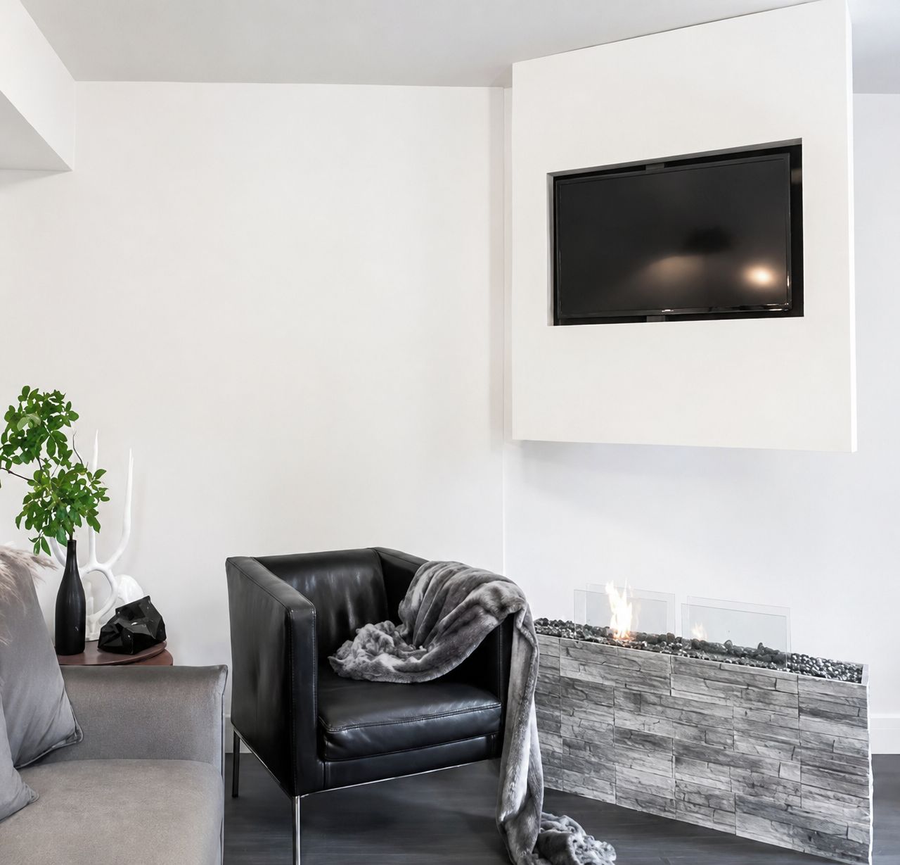 A modern living room with a black leather chair, a grey sofa, a flat-screen TV mounted on the wall, and a stone fireplace.