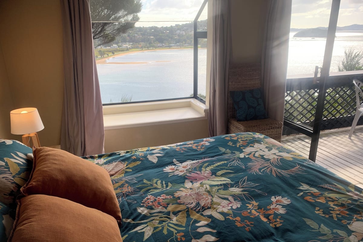 Ein Schlafzimmer mit Bett, Nachttisch, Lampe, Fenster mit Blick auf einen See und Hügel sowie ein Balkon mit Stuhl.