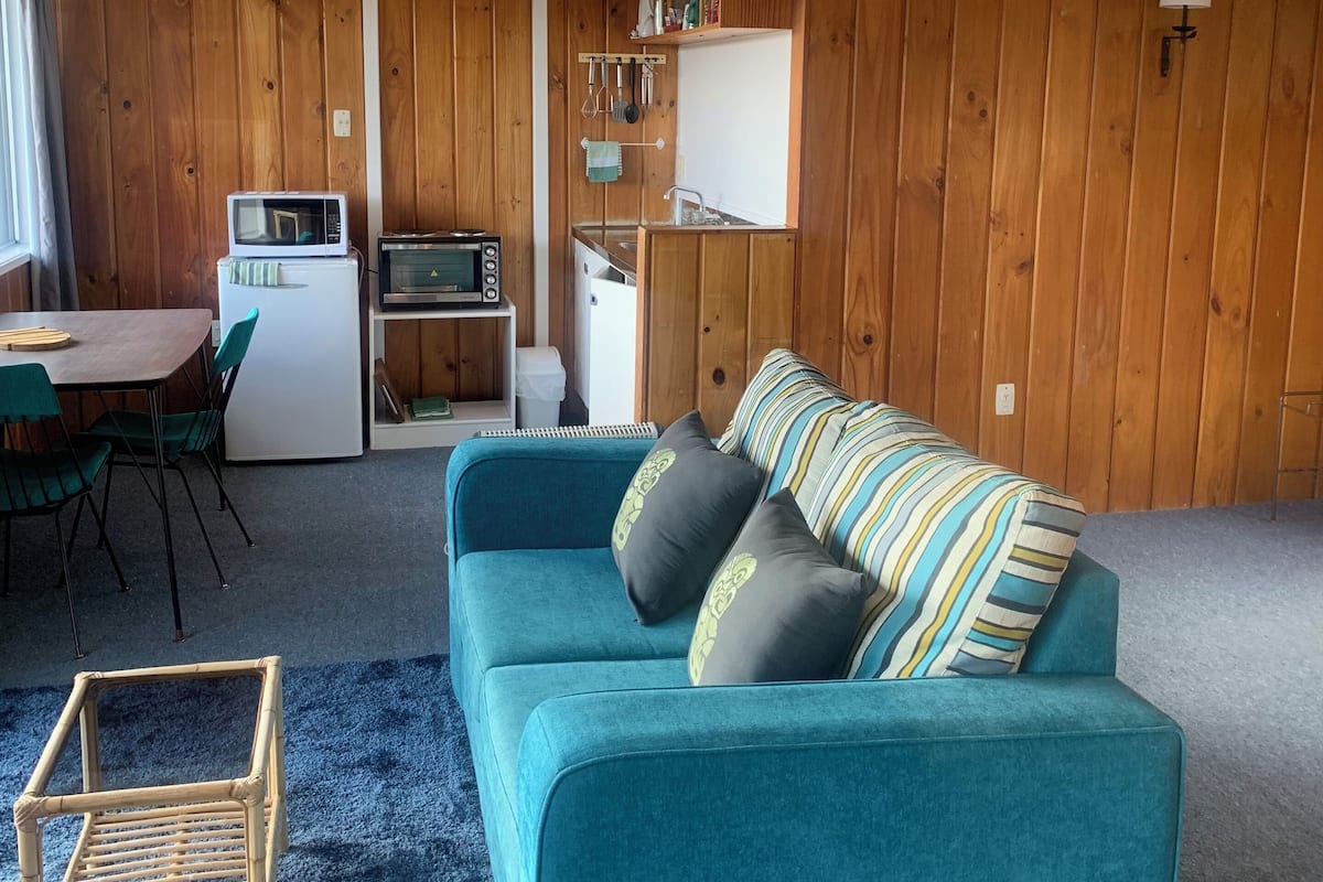 Ein gemütliches Wohnzimmer mit einem türkisfarbenen Sofa, einem kleinen Esstisch und einer Küchenzeile mit Holzwänden und modernen Geräten.