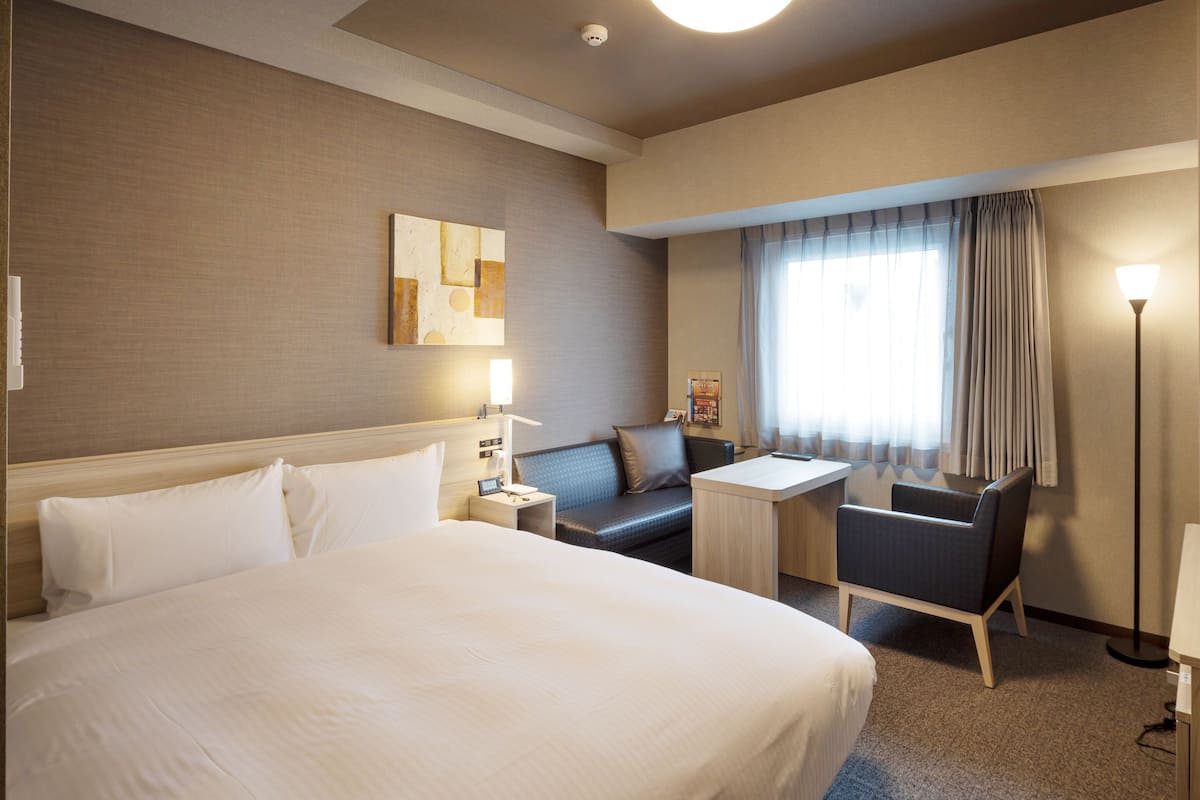 A hotel room with a large bed, a desk, a chair, a sofa, and a lamp.