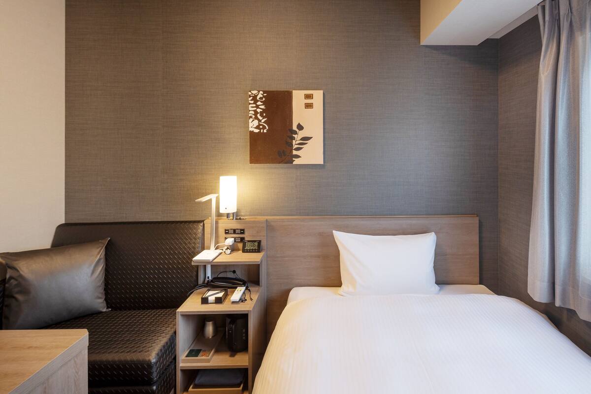 A hotel room with a bed, a desk with a lamp, a chair, and a nightstand with a phone and a clock.