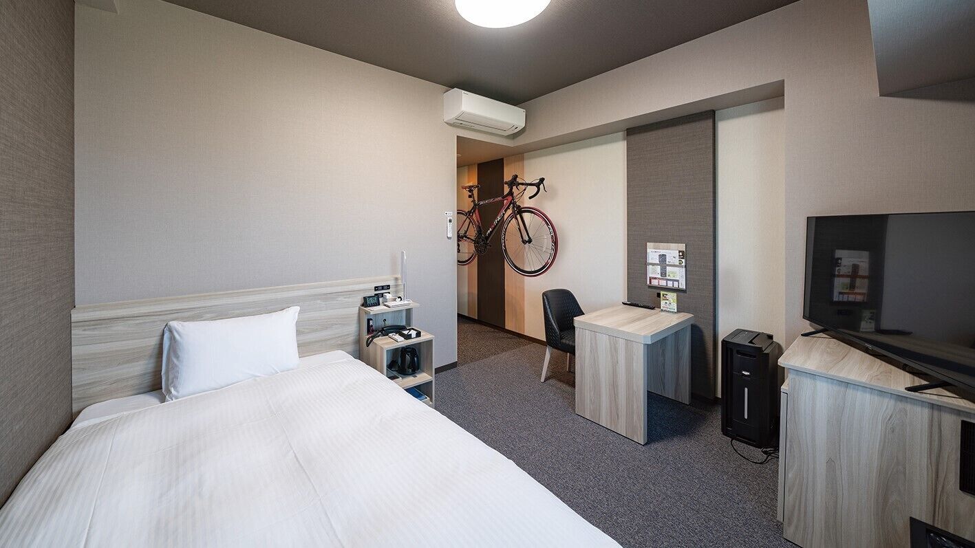 A hotel room with a bed, a desk, a chair, a TV, a bike rack, and a minibar.