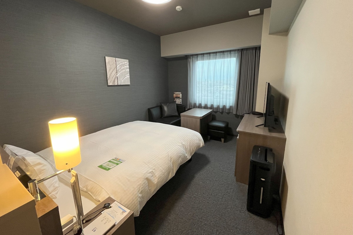 Una habitación de hotel con cama, escritorio, silla, lámpara, televisor y ventana con cortinas.