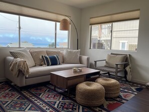 Een gezellige woonkamer met een bank, fauteuil, poufs, een salontafel en een vloerkleed met een patroon.