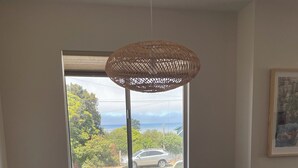 Un coin repas avec une table en bois, deux chaises, un luminaire suspendu en fibres tressées et une fenêtre donnant sur un paysage verdoyant et une voiture.