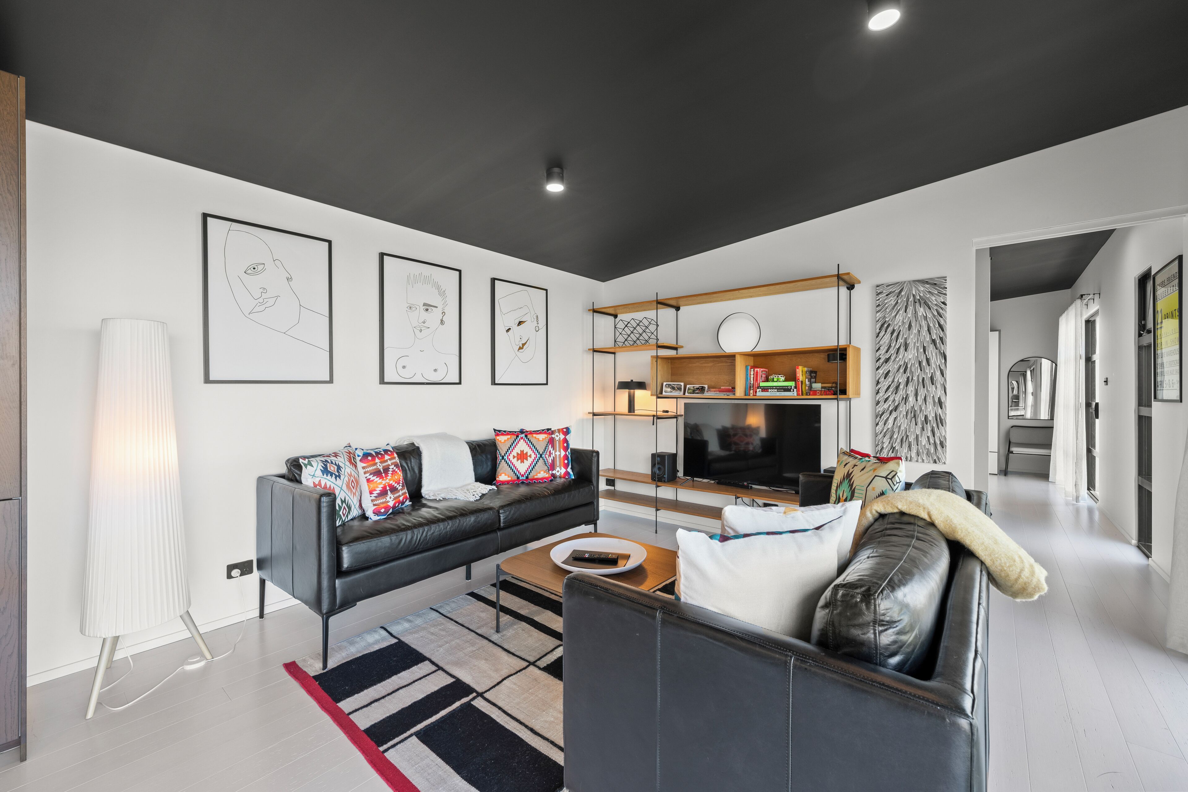 Un salon moderne avec un canapé en cuir noir, une table basse et un téléviseur à écran plat. Des œuvres d’art encadrées ornent le mur et une lampe se trouve dans un coin.