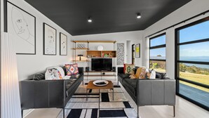 A modern living room with two sofas, a coffee table, and a TV. There are framed artworks on the walls and a large window with a view. - The Outlook on Mount Louis – 1 Bedroom (Tinderbox)