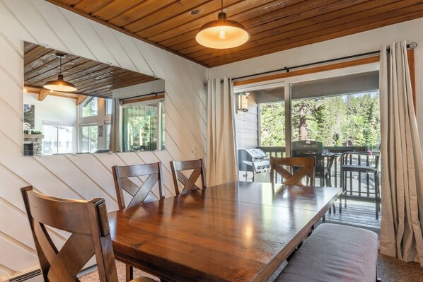 Uma área de refeições com mesa e cadeiras de madeira, janelas amplas e vista para o exterior.