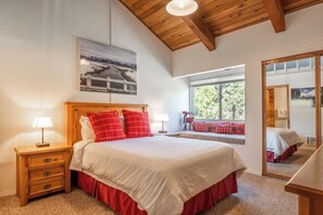 Quarto com cabeceira de madeira, uma cama grande com roupa de cama vermelha e branca, mesa de cabeceira com abajur, janela com vista para vegetação e uma moldura com um quadro na parede.