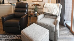 Ein Wohnzimmer mit einem schwarzen Ledersessel, einem grauen Sessel und einem grauen Hocker. Eine Deckenleuchte hängt von der Decke, esstisch mit Stühlen und im Hintergrund eine Küche.