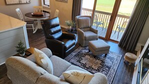 Ein Wohnzimmer mit einer Couch, zwei Sesseln, einem Couchtisch und einem Teppich. Es gibt einen Esstisch mit Stühlen, eine kleine Pflanze und Kunstwerke an den Wänden.