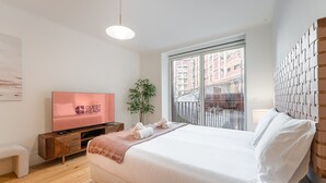 Quarto de hotel moderno com uma cama grande, uma TV em um console de madeira, uma varanda com vista para prédios e uma planta.