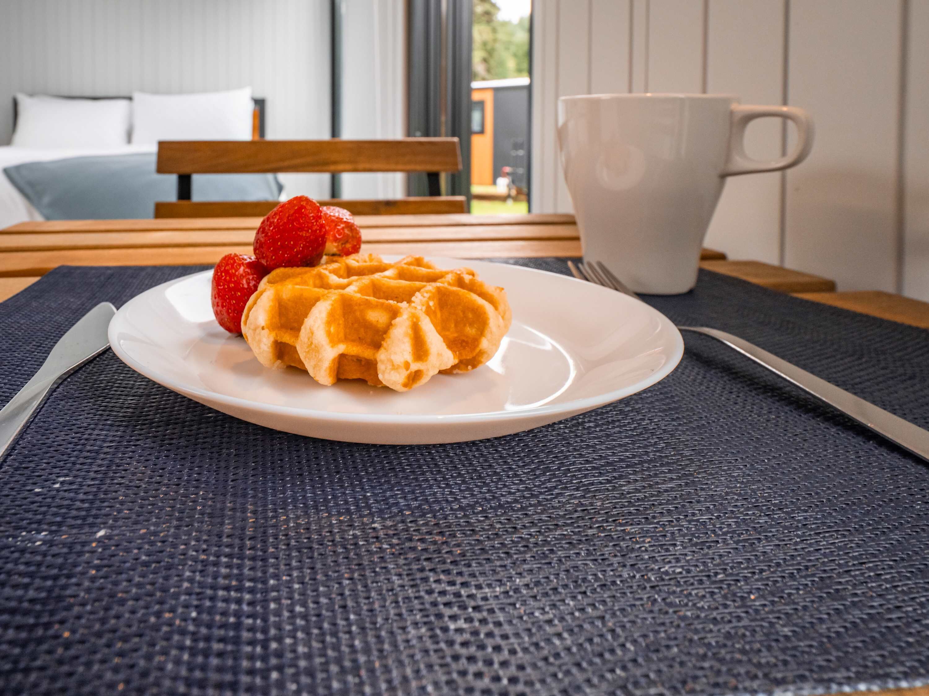 Un plato con gofres y fresas sobre una mesa, una taza blanca y una cama al fondo.