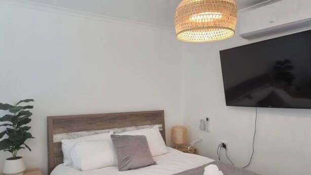 Ein Schlafzimmer mit einem Bett, einem an der Wand montierten Fernseher, einer Topfpflanze und einer gewebten Leuchte.