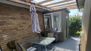 Eine überdachte Terrasse mit einem Glastisch und Stühlen, einem Grill und einer Backsteinmauer.