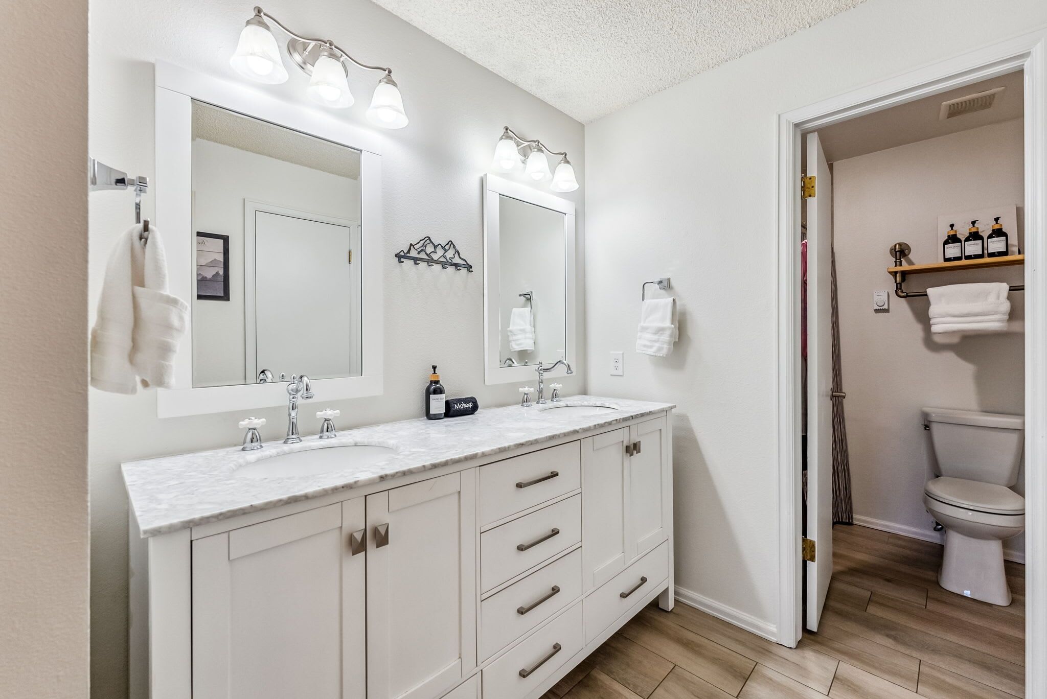 A bathroom with a double sink vanity, a large mirror, and a toilet visible through an open door.