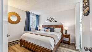 A bedroom with a wooden bed, a round mirror, a wall-mounted geometric decoration, a nightstand with a lamp, and a door with a sign. - Steps to Gondola, Ski-In, 1 Block Main St, Hot Tub (Breckenridge)