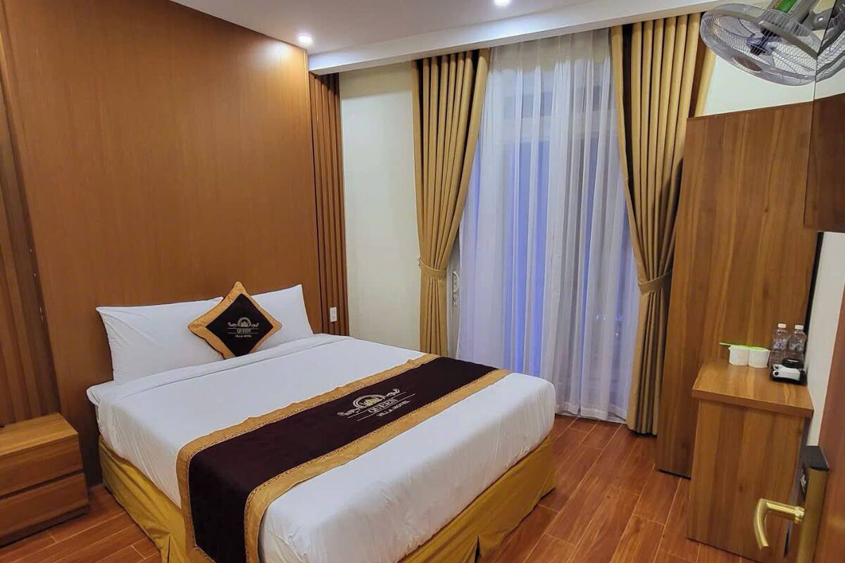 A hotel room with a large bed, wooden flooring, a desk with a chair, a ceiling fan, and a window with curtains.