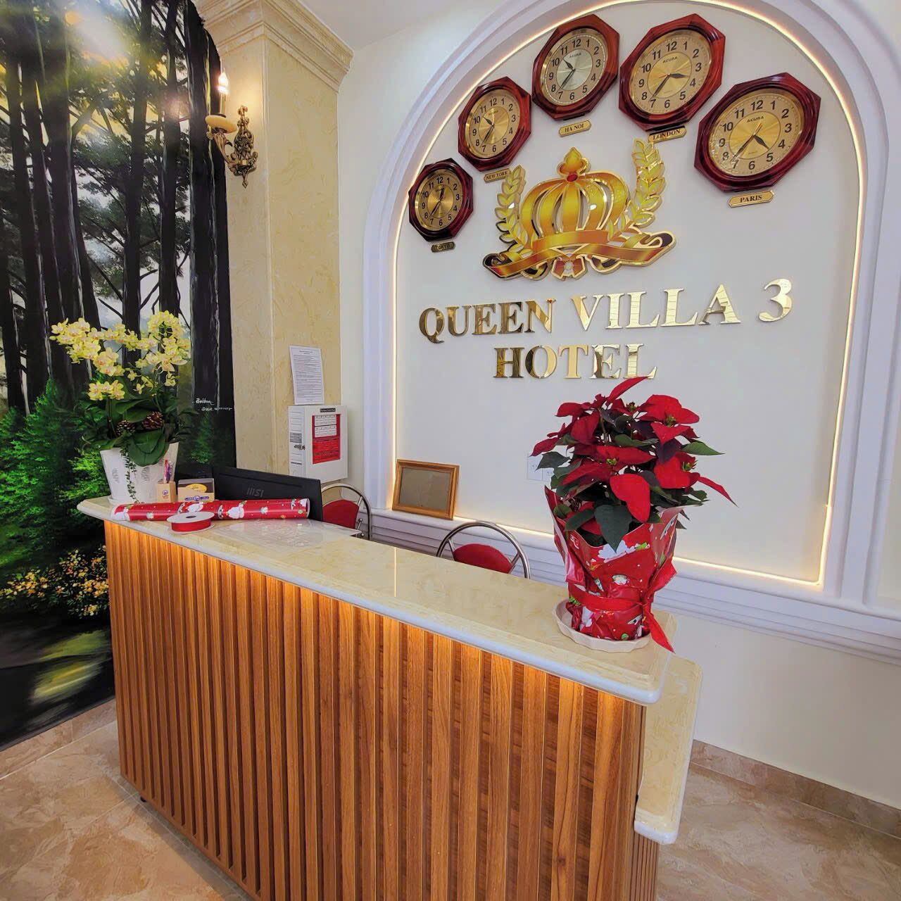 A reception desk at Queen Villa 3 Hotel with a wooden finish, a red vase with red flowers, and multiple clocks on the wall displaying different times.