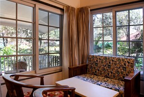Una habitación con sofá, una mesa y dos sillas. Amplias ventanas con cortinas.