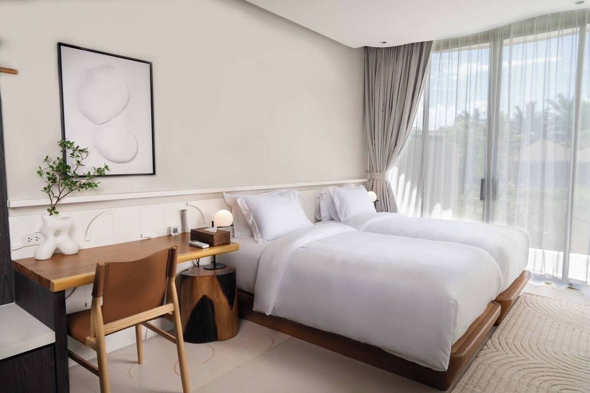 A modern hotel room with a large bed, a desk with a chair, a bedside table, and a large window with curtains.