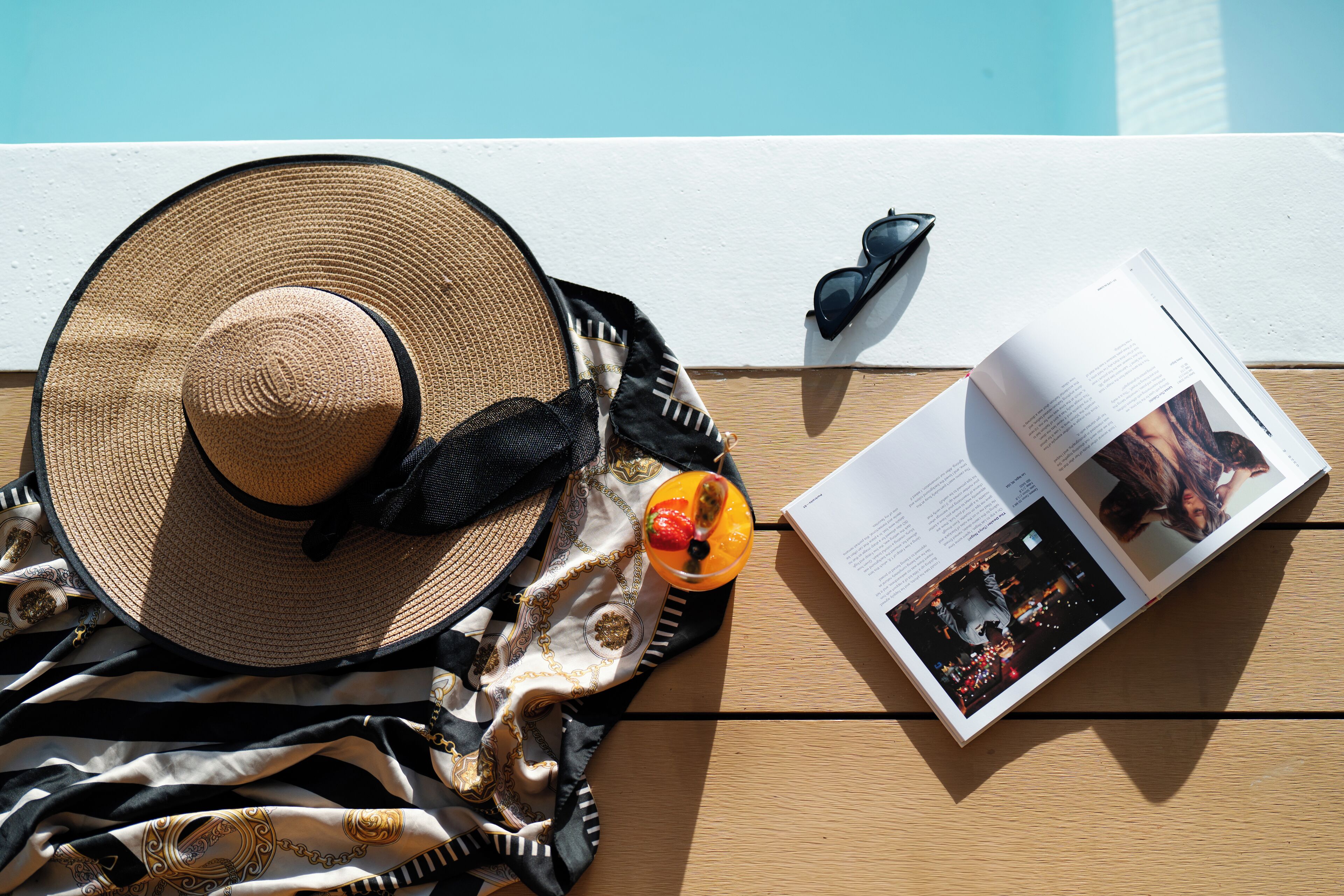 A book, sunglasses, a hat, and a drink on a wooden surface.