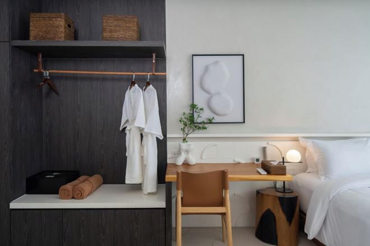 A modern hotel room with a wooden desk, a chair, a bedside table with a lamp, and a bed. There are towels and a plant on the wall.