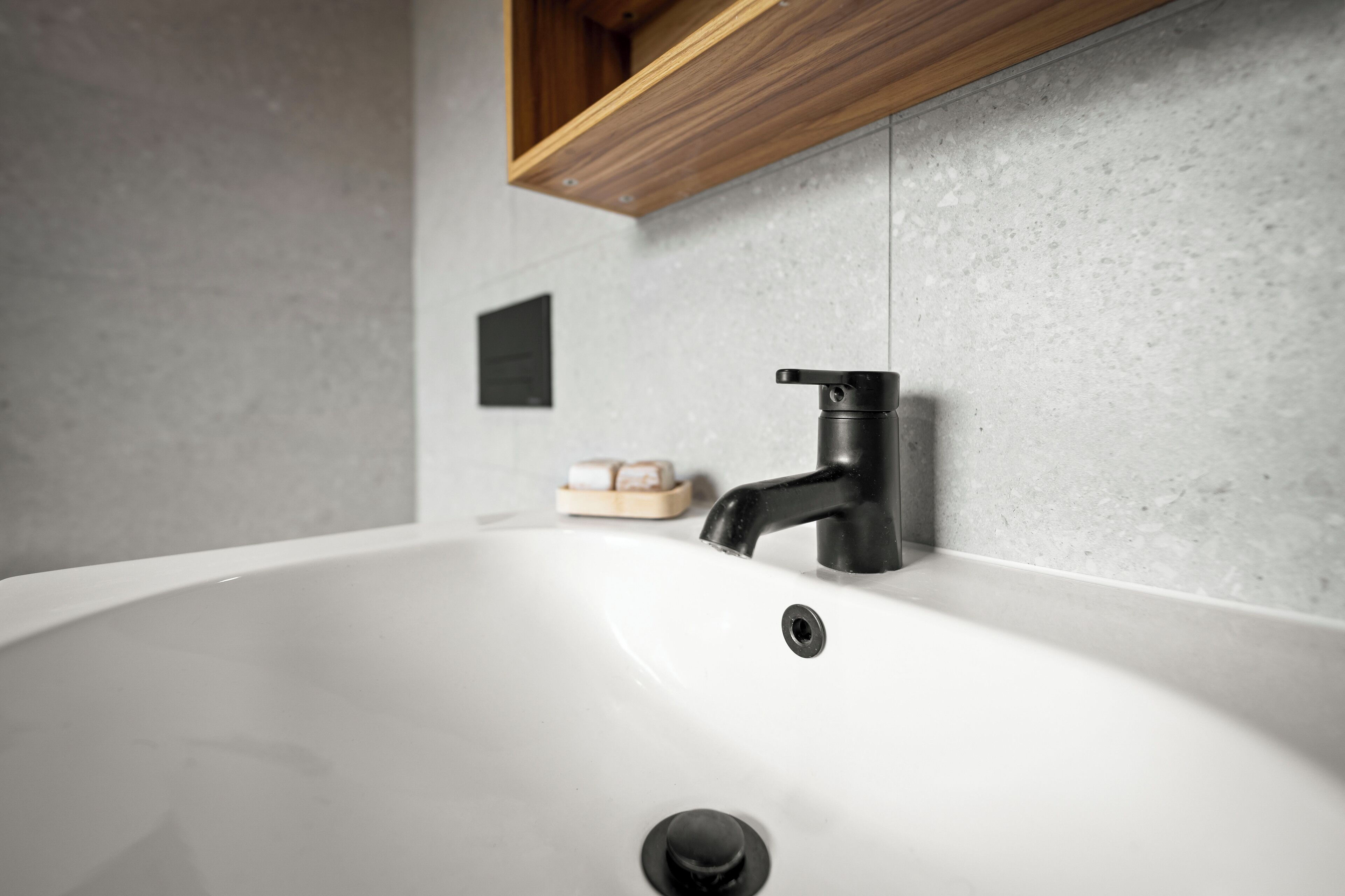 A modern bathroom with a white sink, black faucet, and a wooden shelf above.