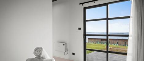 Habitación de hotel moderna con un amplio ventanal corredero a un balcón, una cama bien hecha y un teléfono montado en la pared.