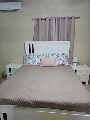 Un lit avec une tête de lit matelassée, un coussin à motifs floraux et une lampe de chevet posée sur une table de chevet blanche.